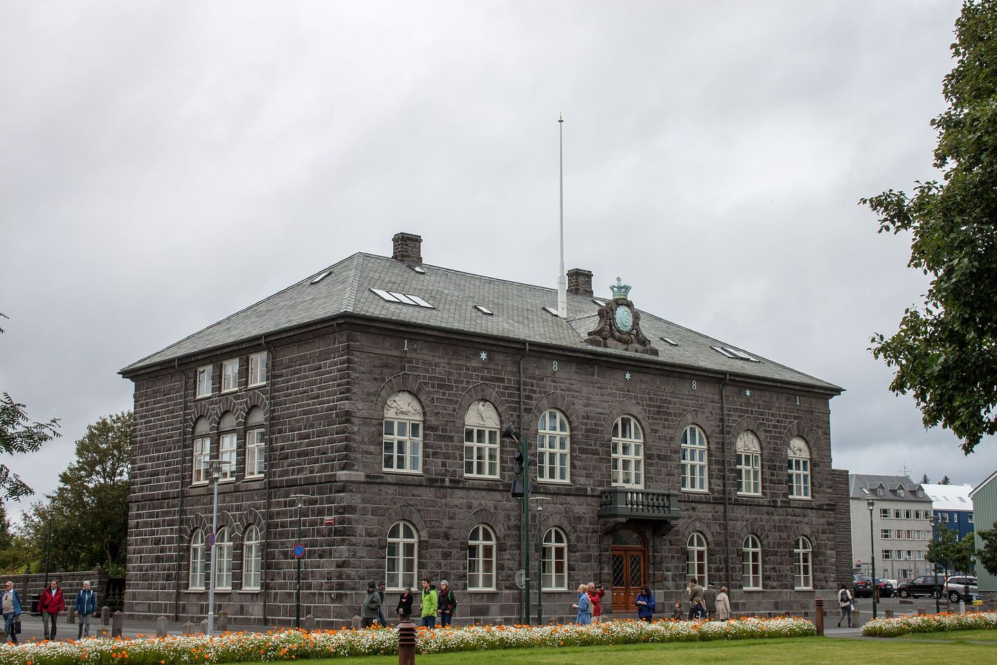 The Alþingi parliament house in Reykjavík, a dark basalt building of 1881 with arched windows, people walking past it on the pavement and a flowerbed in front.