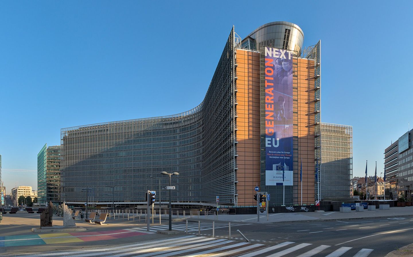 The European Commission's Berlaymont headquarters in Brussels under a clear sky, a Next Generation EU banner hung down its tower, and the crossings in front of it empty.