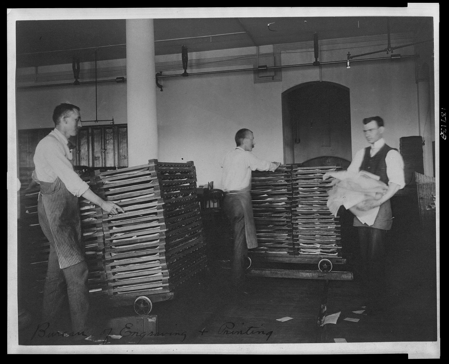 Three men wheeling racks of freshly printed banknote sheets to dry, in an archive photograph of the Bureau of Engraving and Printing.