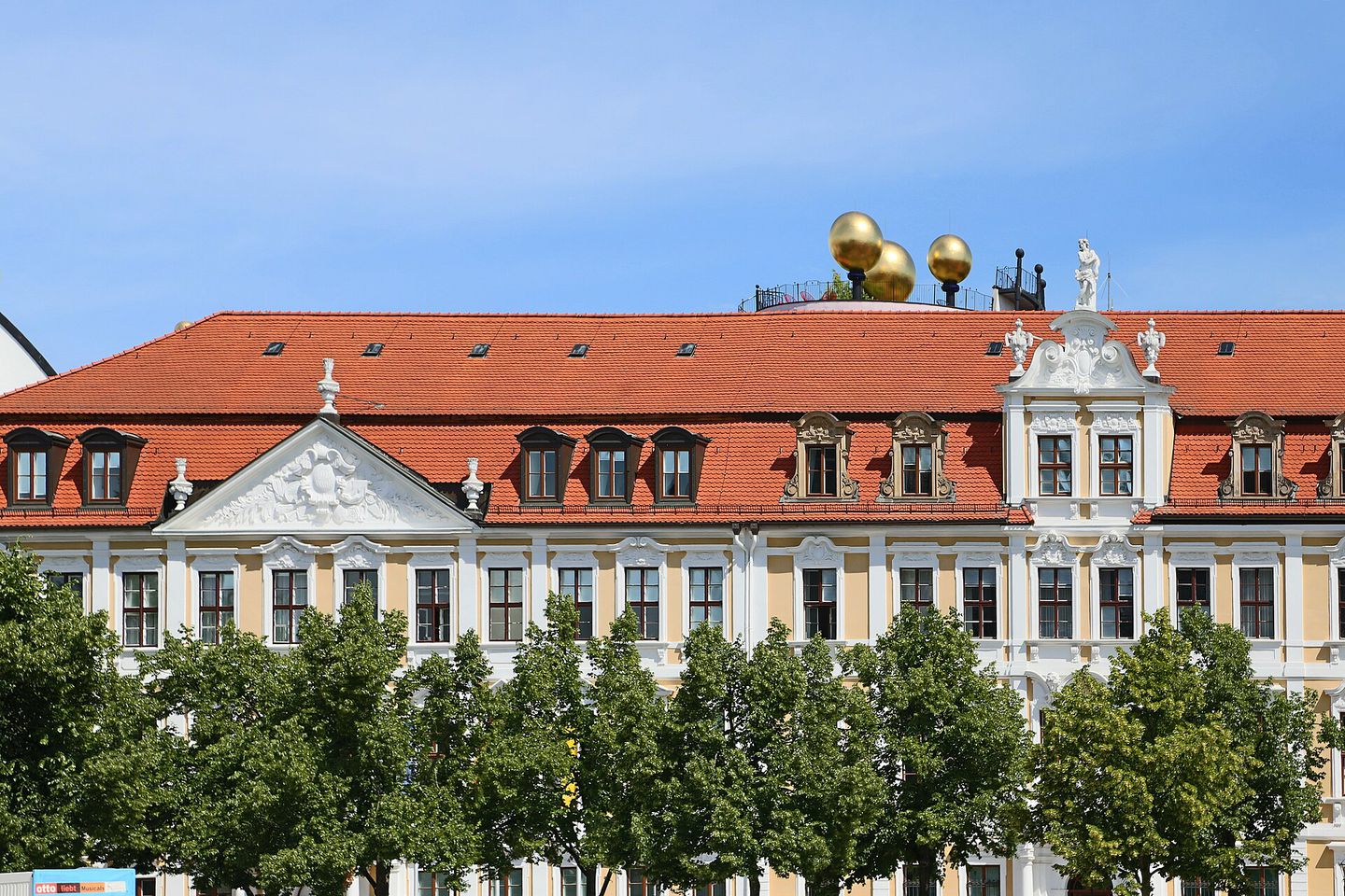 La façade baroque du parlement de Saxe-Anhalt, sur la Domplatz de Magdebourg, son toit rouge surmonté de trois sphères dorées, vue par-dessus une rangée d'arbres.