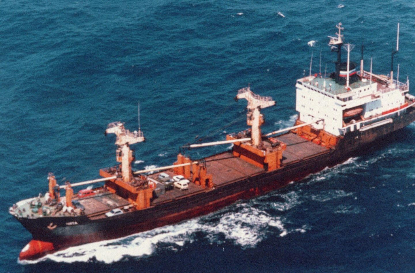A general cargo ship under way at sea, cars stowed on its open deck, seen from the air.
