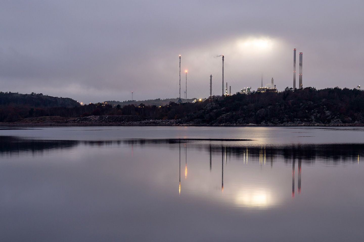 The lights of a coastal oil refinery under fog, reflected across still water.